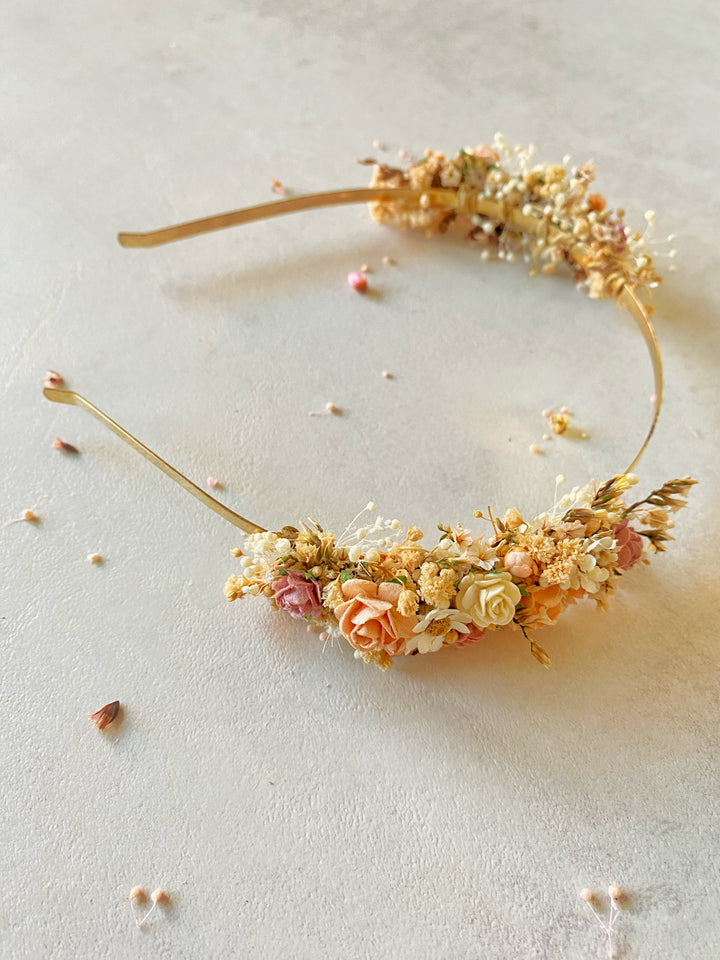 A gold metal headband decorated with dried flowers in soft peach, cream, and muted pink tones, arranged in a curved shape on a light neutral surface.