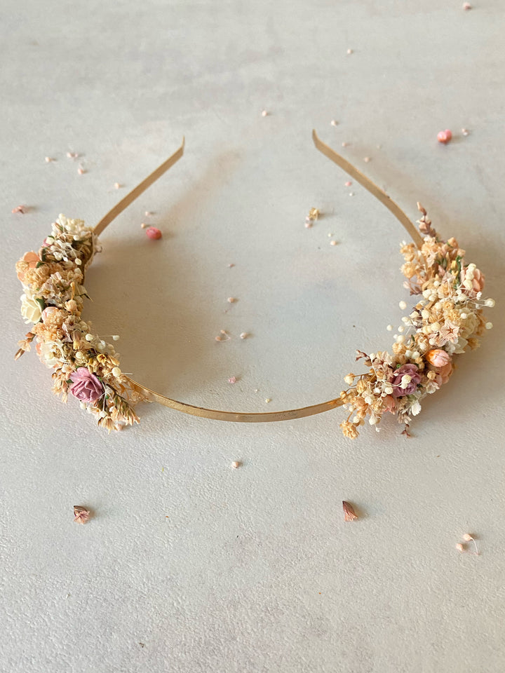 A gold metal headband decorated with flowers and greenery in soft pink, beige, and cream tones, photographed on a light neutral surface with scattered petals around it.