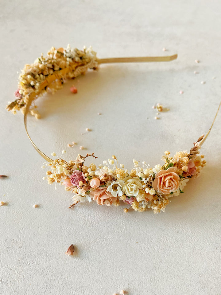 A delicate gold headband decorated with flowers, small white blossoms, and peach-colored roses, displayed on a textured light beige surface.