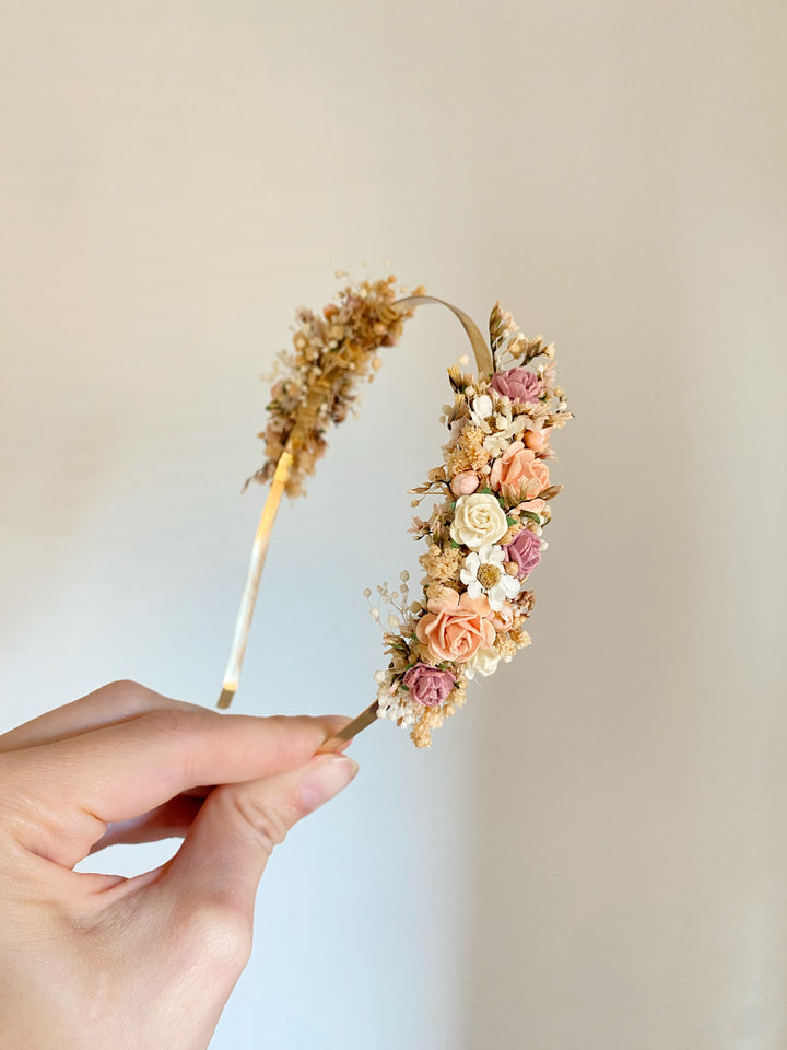 A hand holding a gold metal headband decorated with flowers in soft peach, cream, and dusty pink tones against a plain light beige background.