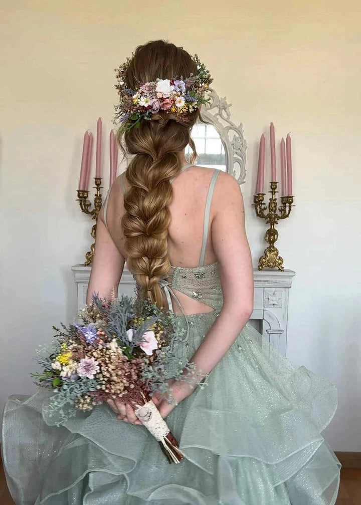 A woman seated with her back to the camera, wearing a voluminous pale sage green gown and holding a pastel floral bouquet. Her hair is styled in a long braid with a floral crown, set against an indoor background with a mirror and pink candlesticks.