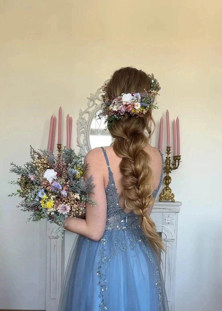 A woman viewed from behind wearing a light blue sleeveless tulle gown with beaded details, holding a large floral bouquet. She has a long braided hair adorned with a flower crown, standing in front of a white fireplace mantel with pink candles.