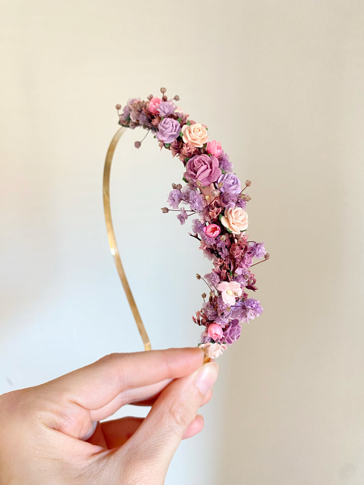 A hand holding a gold metal headband decorated with pink, purple, and peach flowers against a soft beige background.