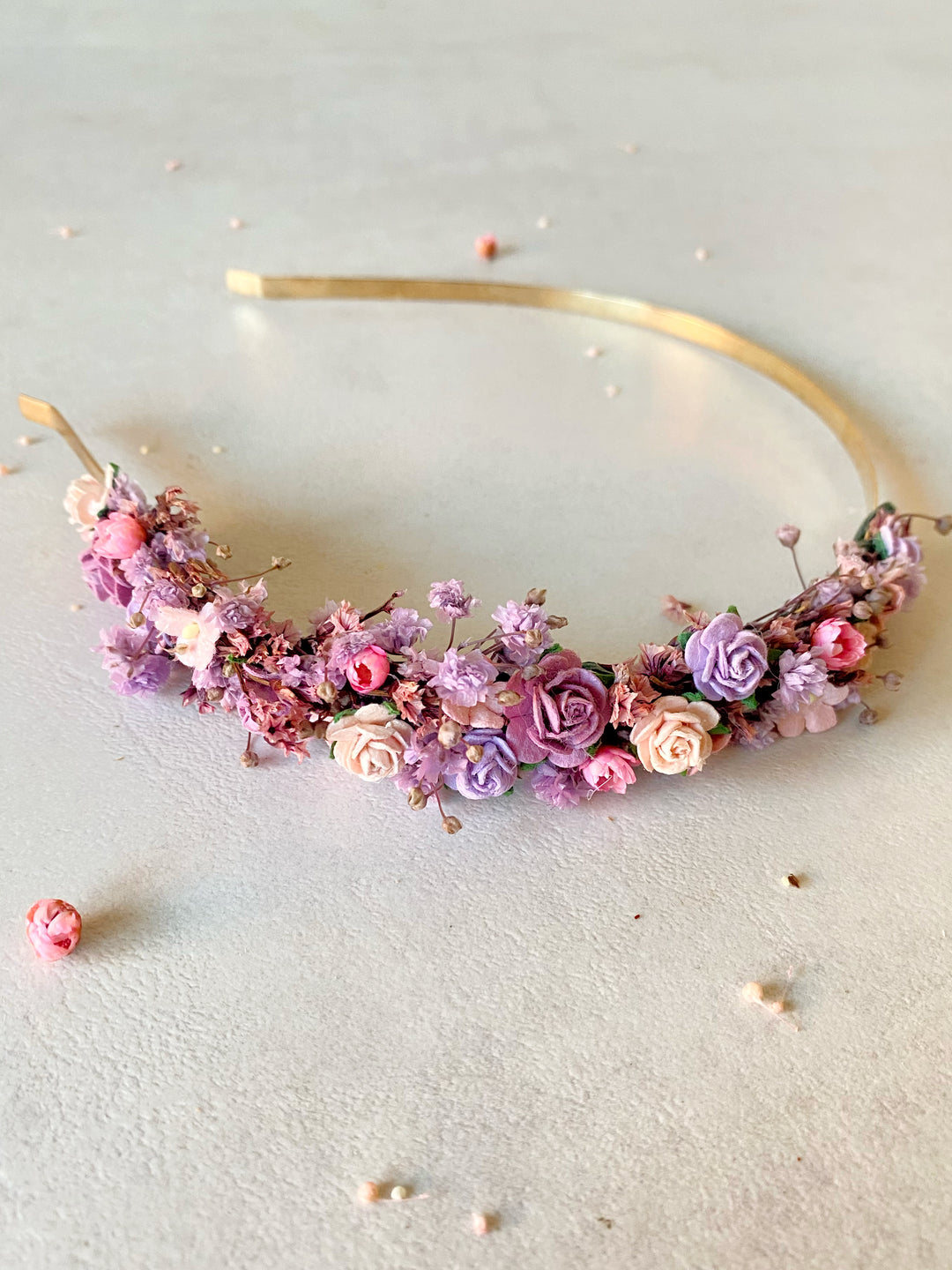 A gold metal headband decorated with a dense cluster of pink, purple, and peach flowers, photographed on a light textured surface.