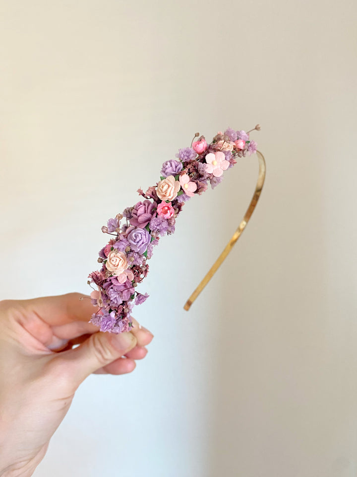 A hand holding a gold headband decorated with small pink and purple flowers against a plain light beige background.