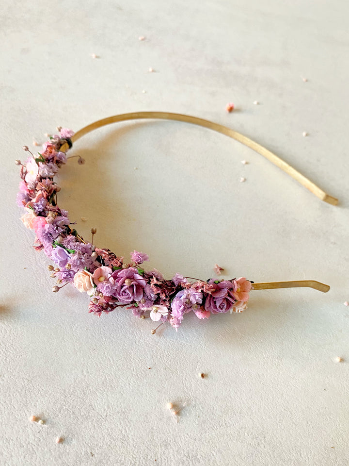 A floral headband with a gold metal band decorated with small pink, purple, and peach dried flowers, photographed on a light textured surface.