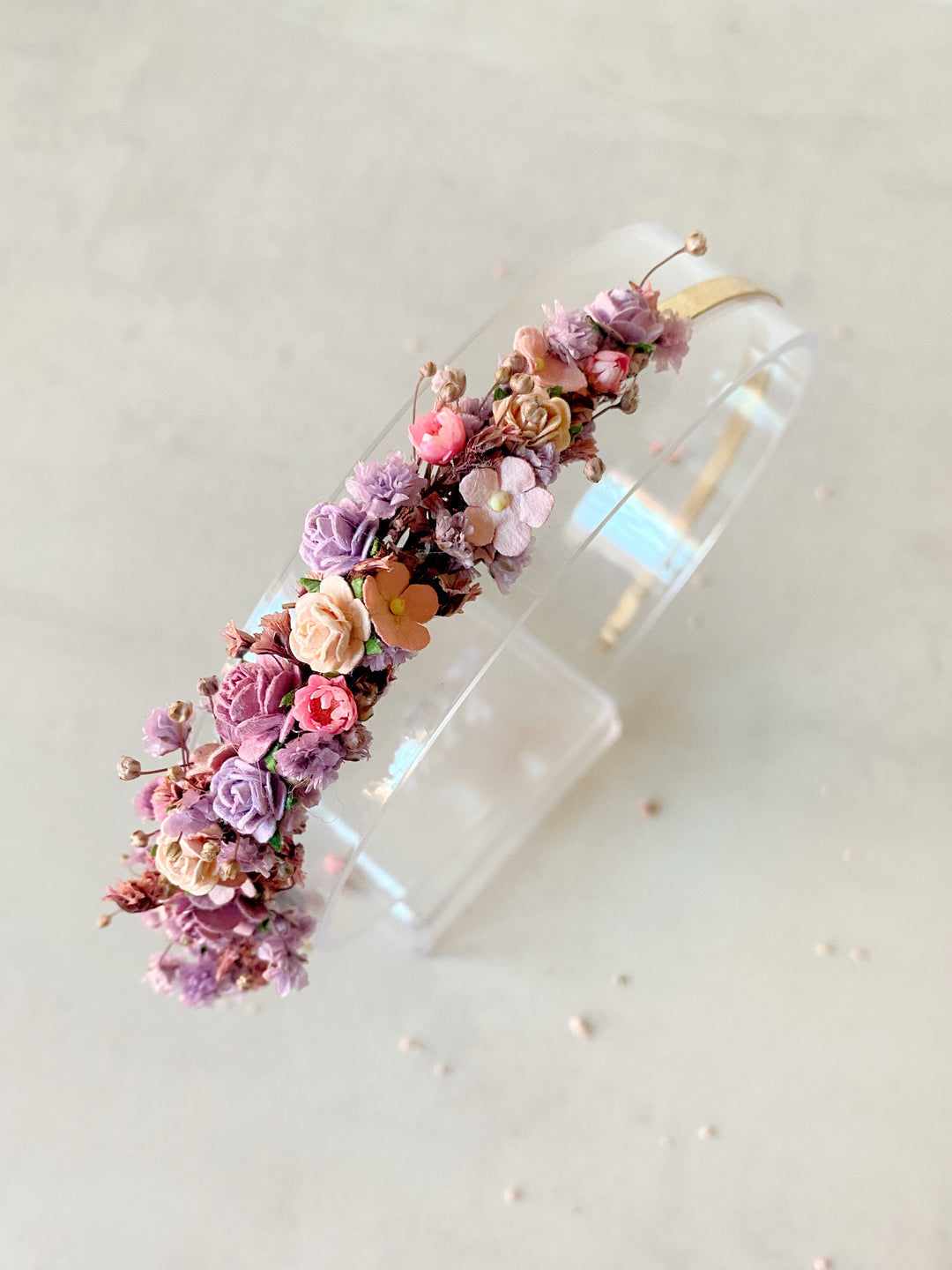 A clear acrylic headband decorated with a dense cluster of pastel flowers in pink, purple, peach, and orange, displayed on a clear stand against a light beige background.