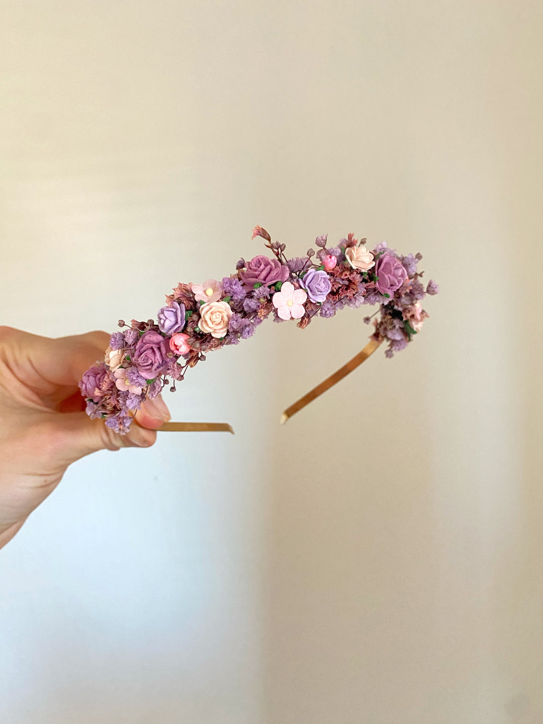 A hand holding a delicate floral headband decorated with small purple, pink, and lavender flowers on a gold band, set against a plain light beige background.
