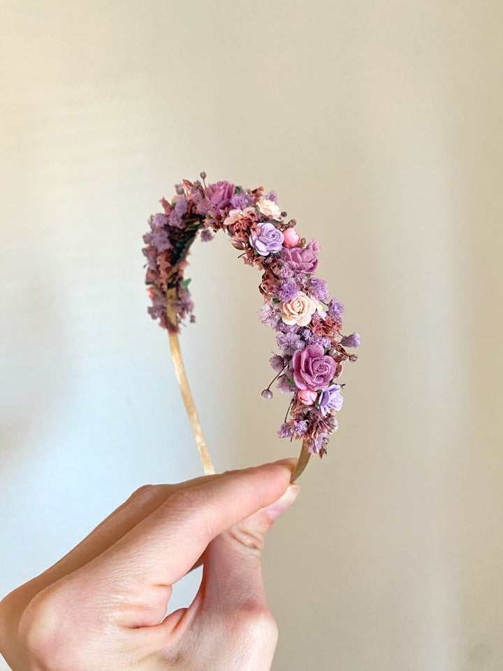 A hand holding a delicate floral headband decorated with small pink, purple, and lavender flowers on a gold-toned band, set against a soft beige background.