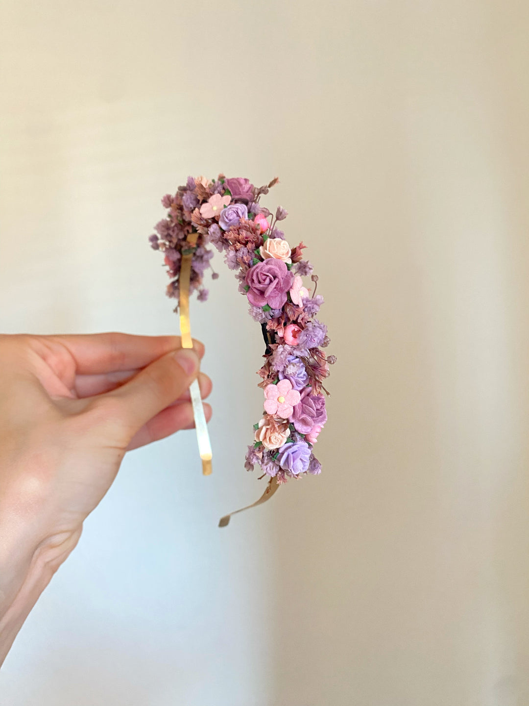 A hand holding a delicate floral headband decorated with small pink, purple, and peach dried flowers on a gold-tone band, set against a plain beige background.