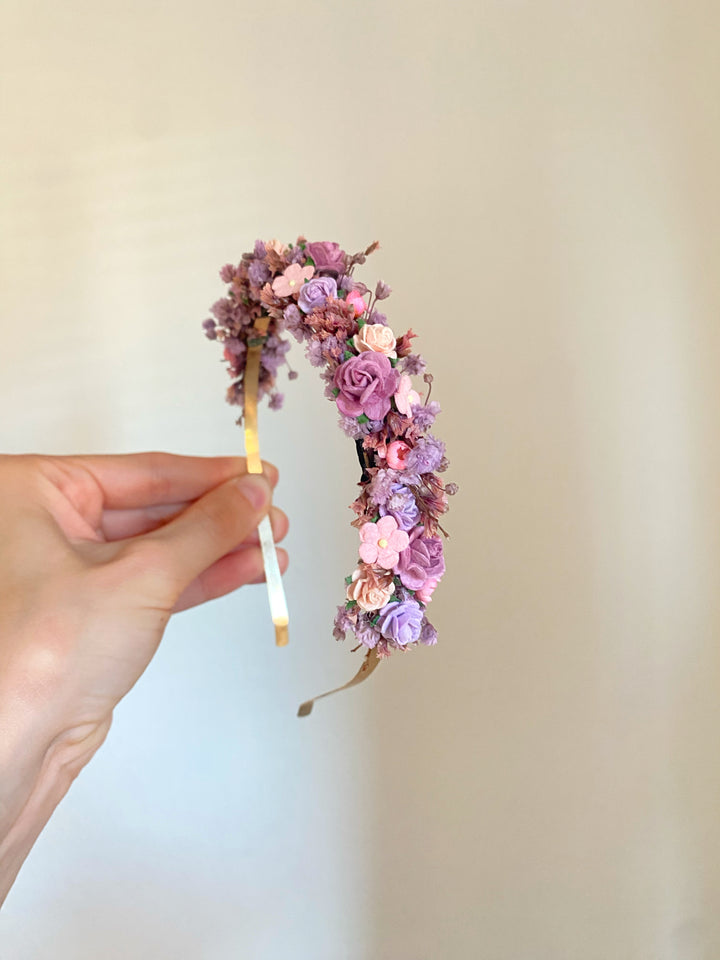 A hand holding a delicate floral headband decorated with small pink, purple, and peach dried flowers on a gold-tone band, set against a plain beige background.
