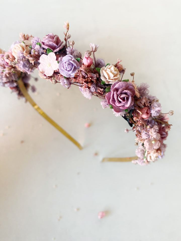 A delicate floral headband featuring pink, purple, and peach flowers with a gold-tone band, photographed on a light cream background.
