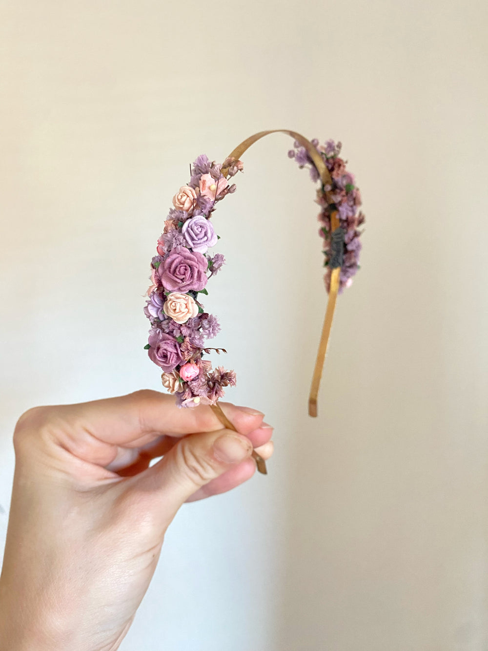 A hand holding a floral headband decorated with pink, purple, and peach flowers on a light beige background.