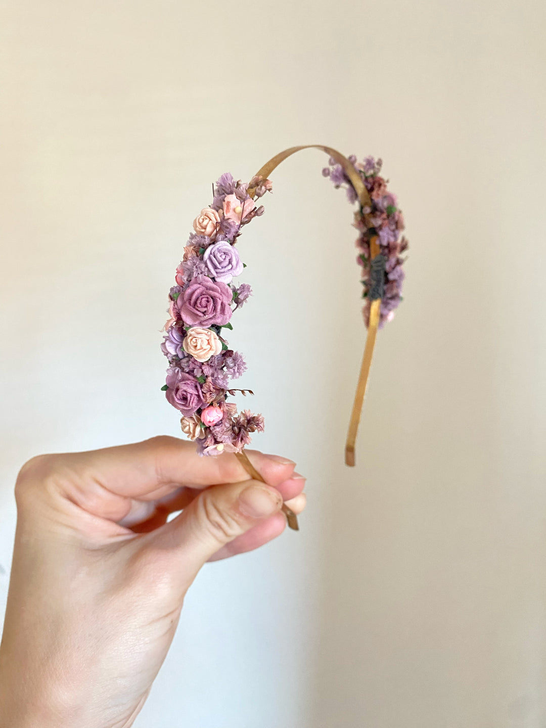 A hand holding a floral headband decorated with pink, purple, and peach flowers on a light beige background.