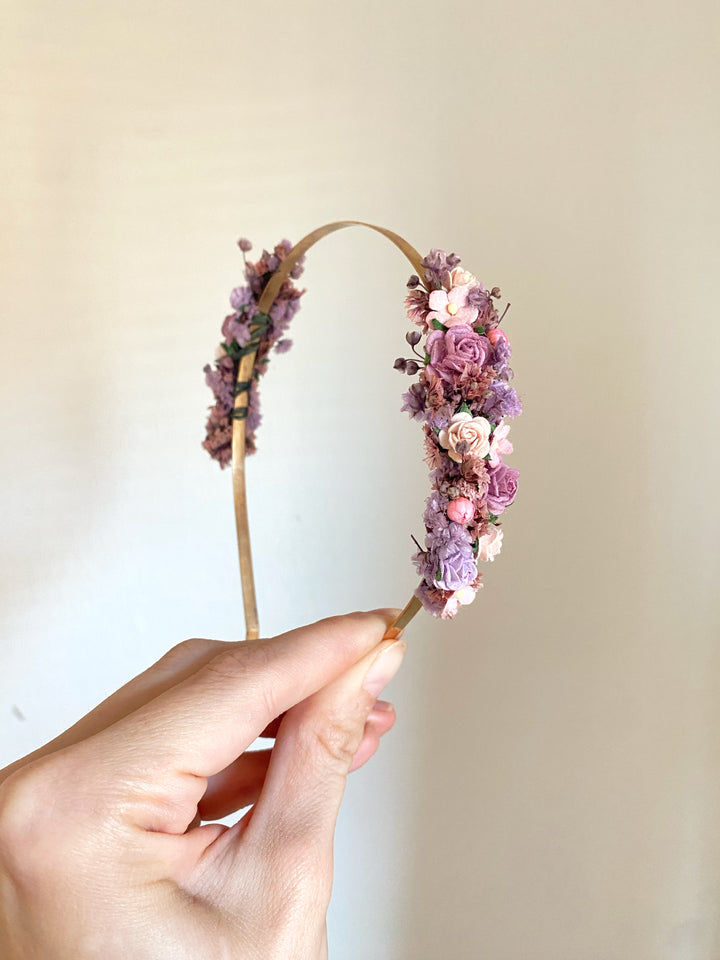 A hand holding a thin gold headband decorated with pink, purple, and lavender flowers against a plain beige wall.