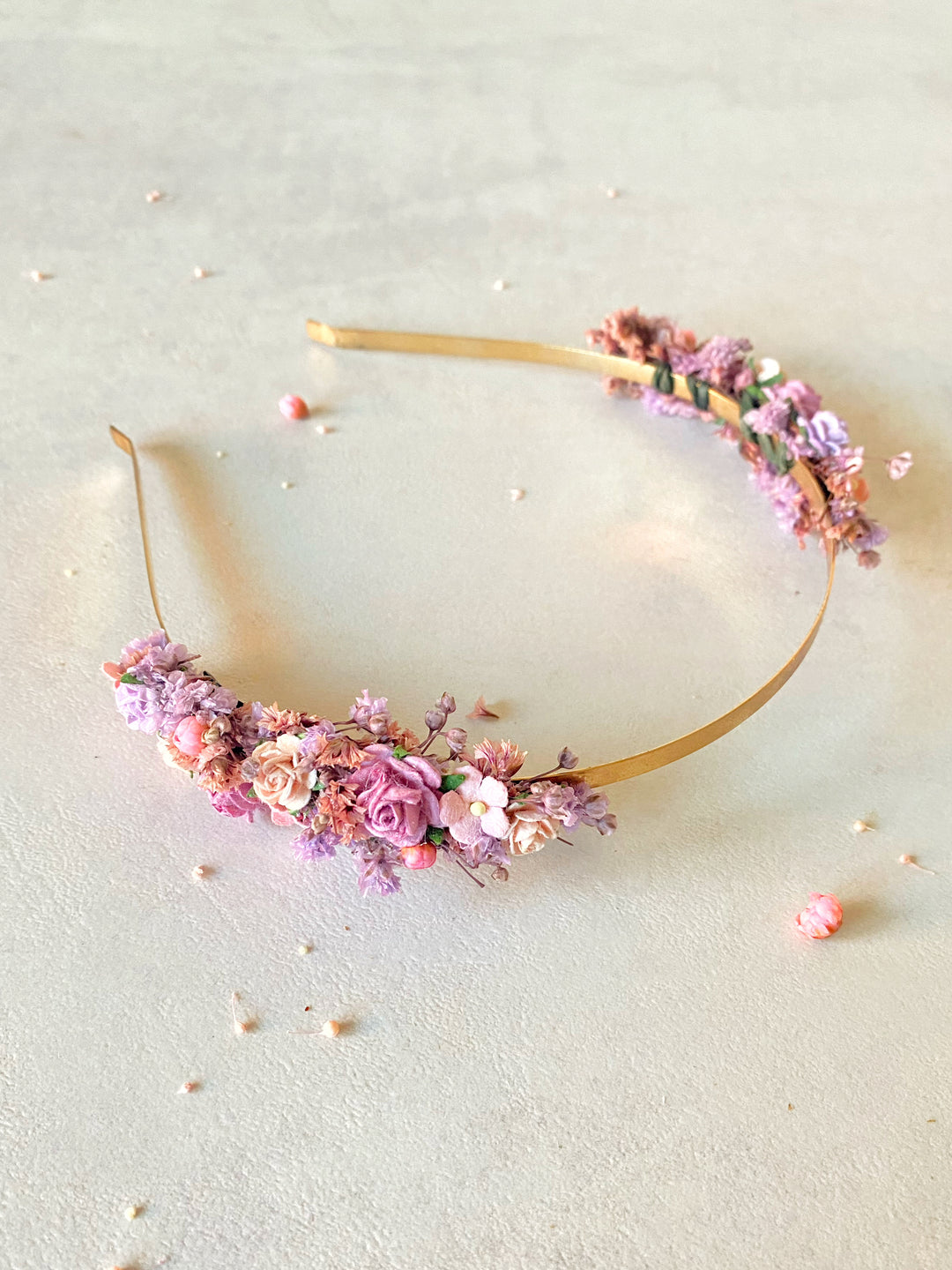 A floral headband with a gold band and clusters of pink, purple, and peach flowers, displayed on a textured beige surface.