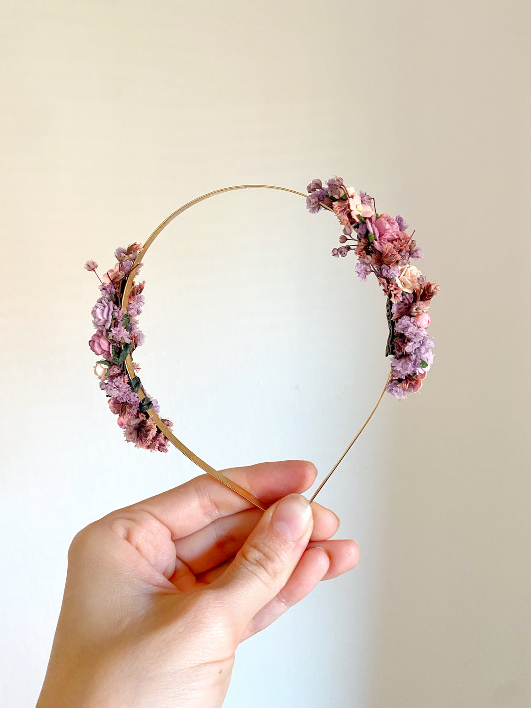 A hand holding a thin gold wire headband decorated with pink and purple flowers against a plain light beige background.