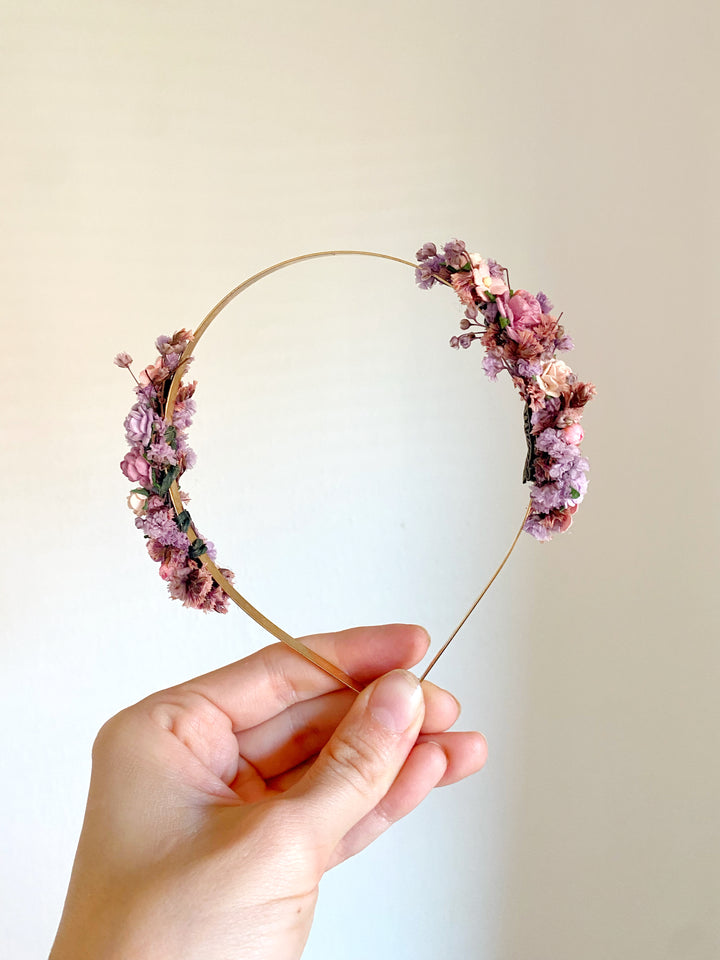 A hand holding a thin gold wire headband decorated with pink and purple flowers against a plain light beige background.