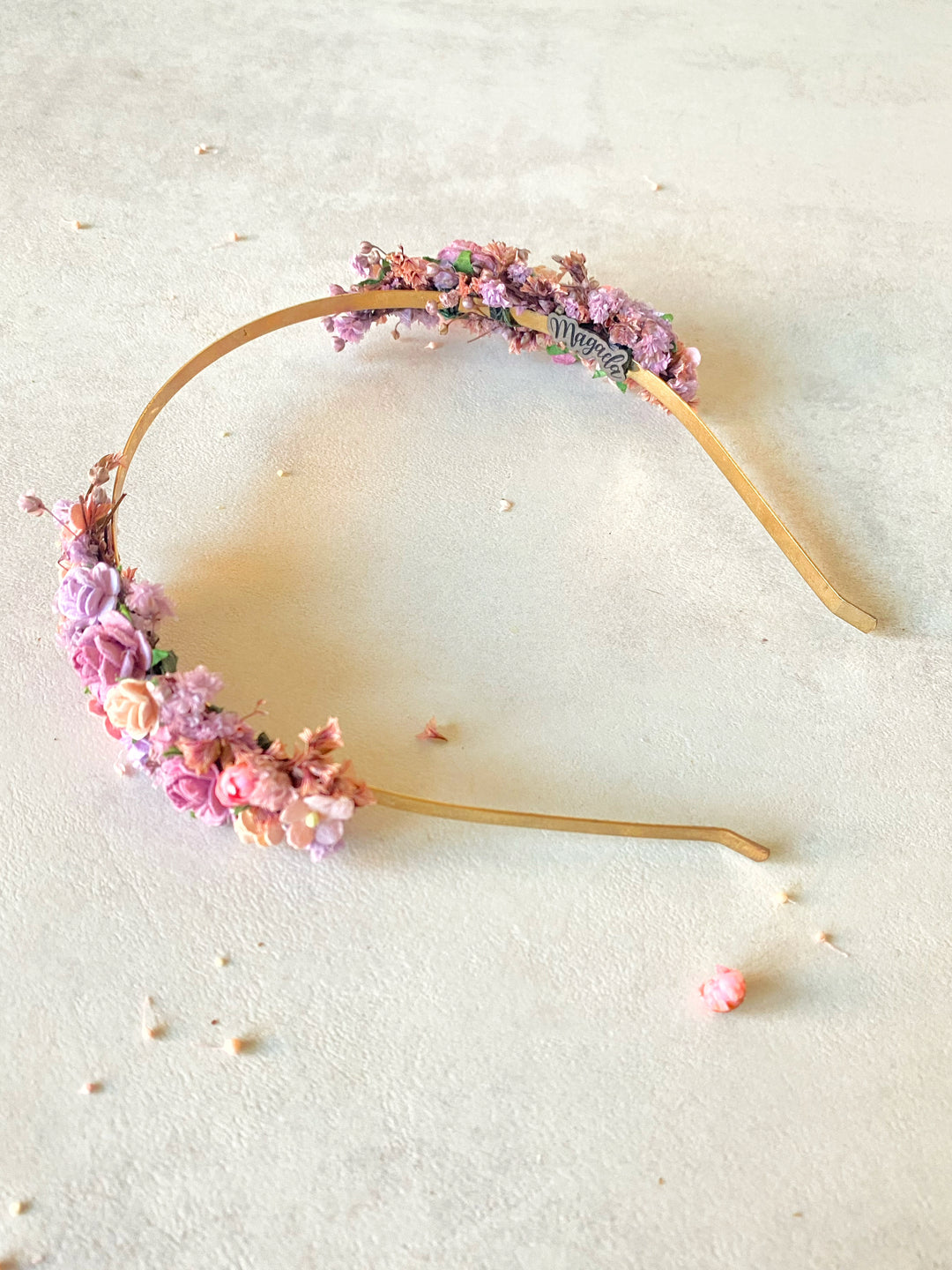 A floral headband with pink and purple flowers and green leaves, displayed on a textured beige surface.