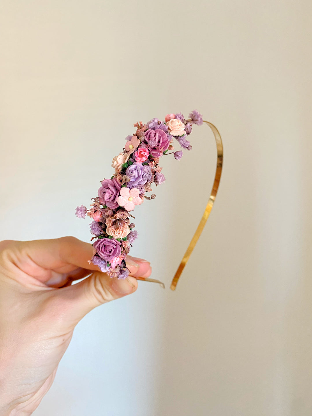 A hand holds a gold metal headband decorated with small pink, purple, and mauve flowers against a plain light beige background.
