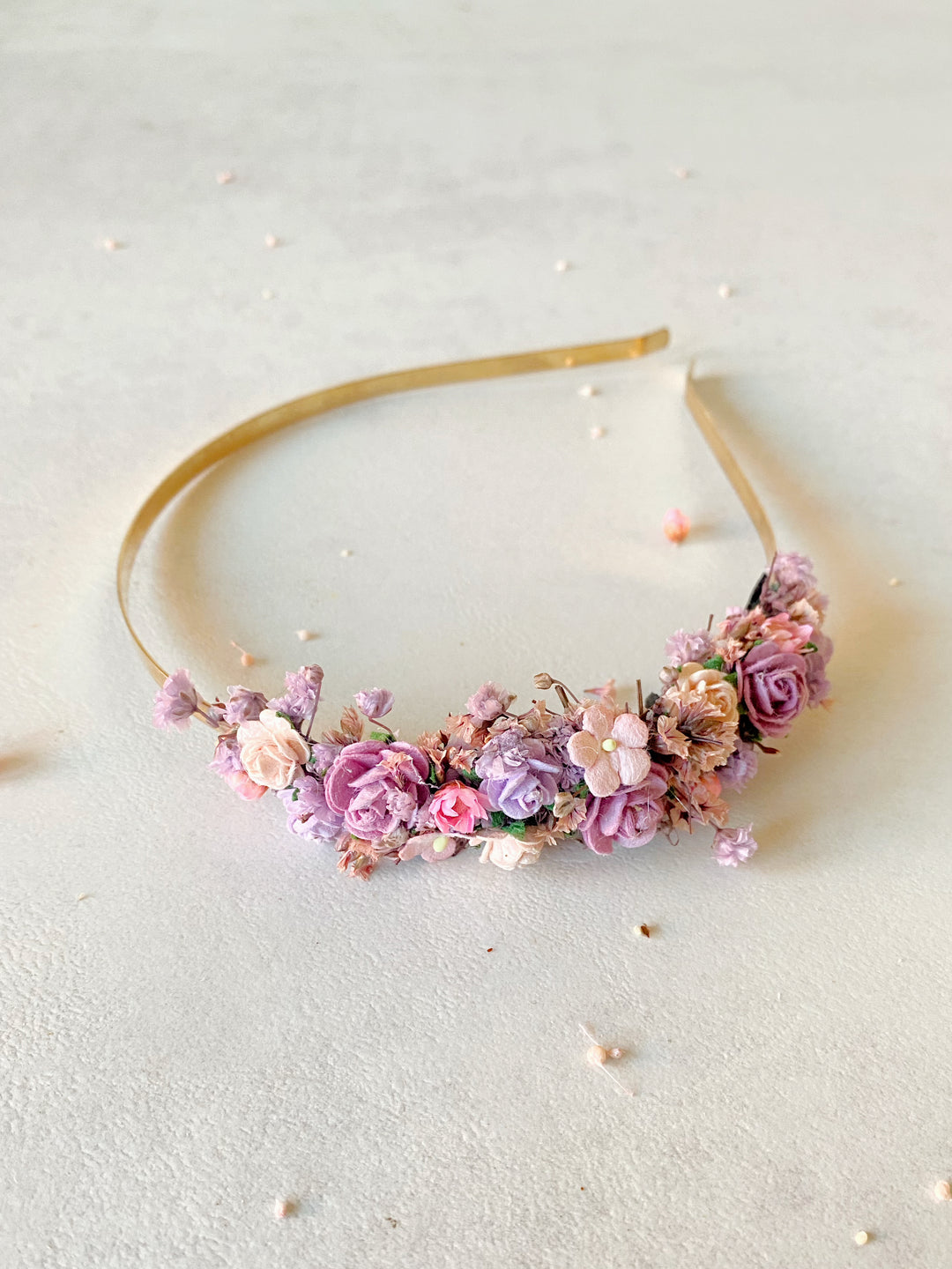 A gold metal headband decorated with a dense cluster of small artificial and preserved  flowers in shades of pink, lavender, and peach, photographed on a light neutral textured surface.