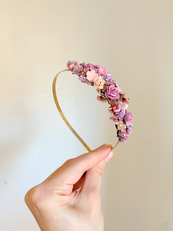 A hand holding a gold metal headband decorated with small pink, purple, and peach flowers against a plain light beige background.