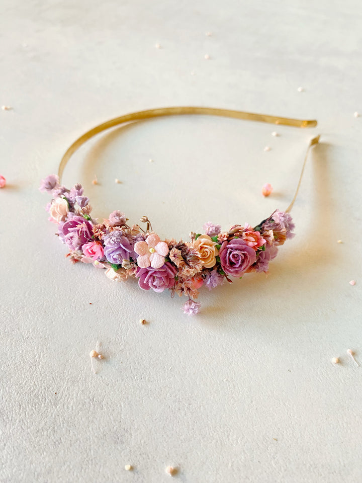 A delicate floral headband featuring a curved arrangement of pink, purple, peach, and white flowers on a thin gold band, photographed on a light textured surface with scattered petals.