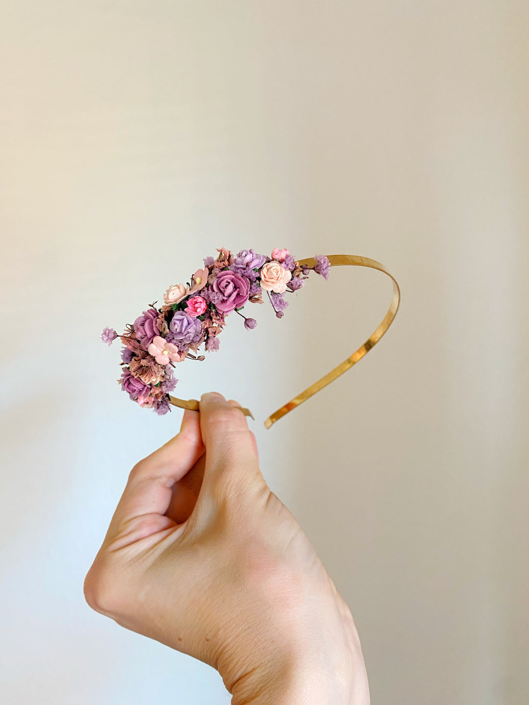 A hand holding a gold headband decorated with pink and purple flowers against a plain beige background.