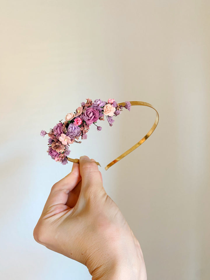 A hand holding a gold headband decorated with pink and purple flowers against a plain beige background.