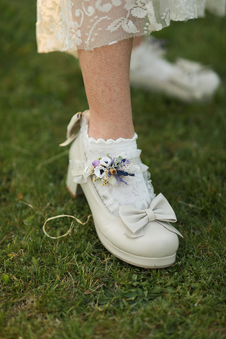 White lace-up shoes with decorative flower shoe clip in purple tones and bows on a grassy background
