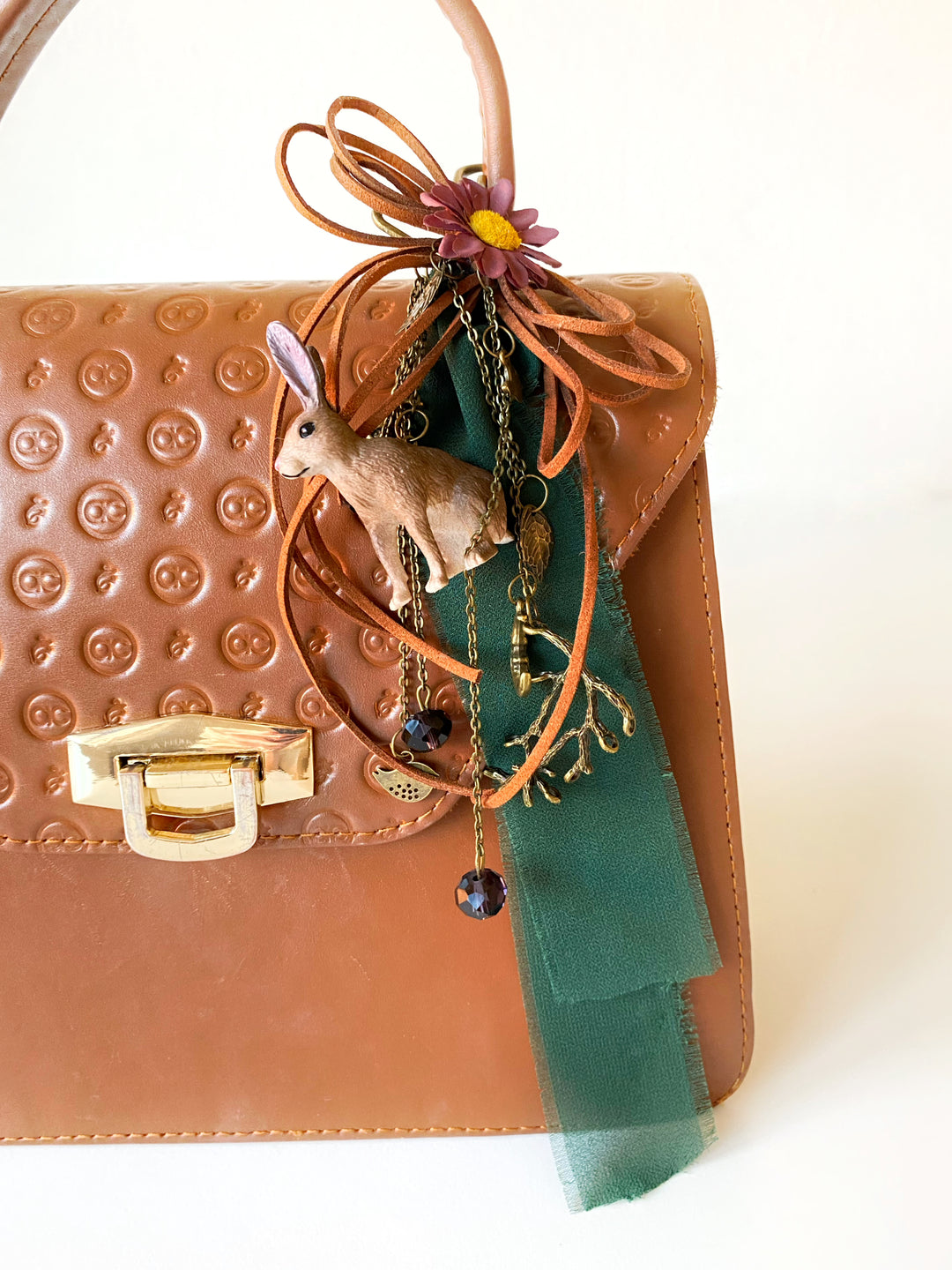 A close-up of a brown textured leather handbag with a gold-tone clasp, decorated with a woodland rabbit -themed hanging charm featuring a small rabbit figurine, a green ribbon, and a purple flower.