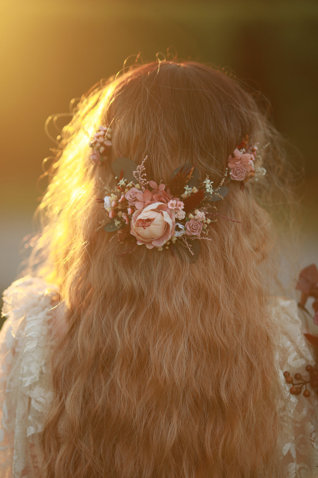 A close-up back view of a person with long wavy light brown hair wearing a floral hair accessory with pink and cream flowers and green leaves, set against a warm golden outdoor background.