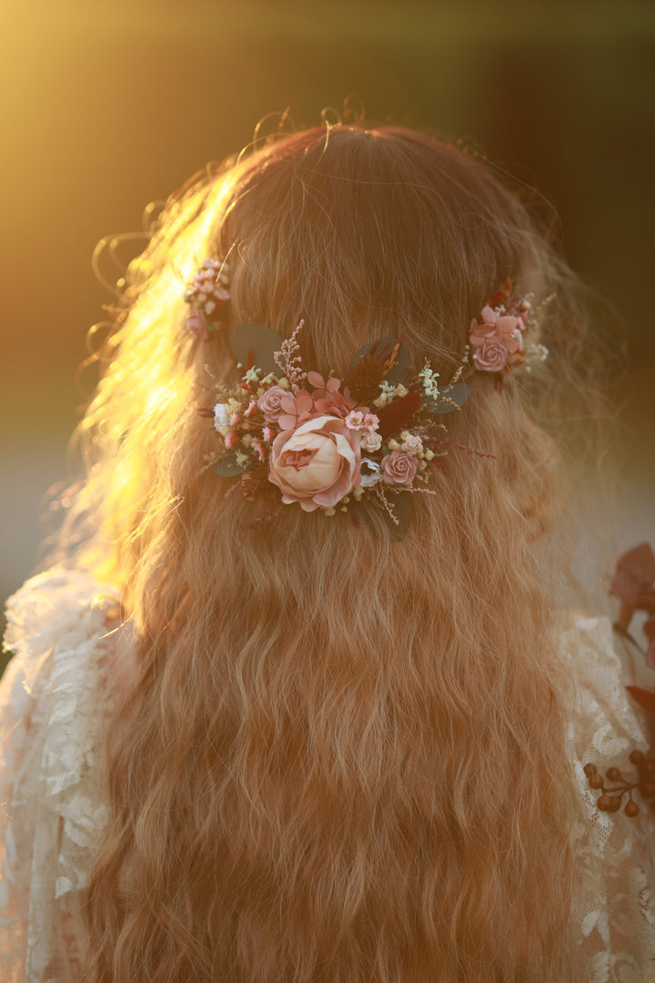 A close-up back view of a person with long wavy light brown hair wearing a floral hair accessory with pink and cream flowers and green leaves, set against a warm golden outdoor background.