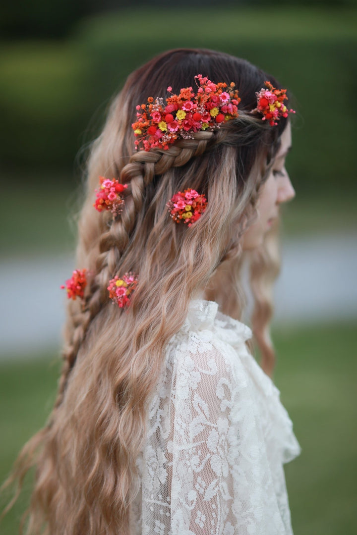 A woman in profile wearing a long blonde braid adorned with bright red and orange floral hair accessories, set against a soft blurred green outdoor background.
