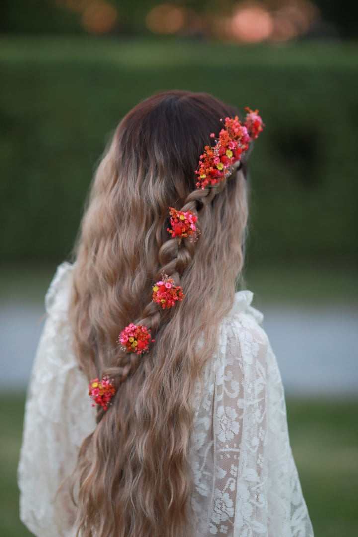 Long wavy blonde-to-brown ombre hair styled in a side braid with bright orange and red flower hair accessories, shown from the back on a person wearing a white lace top outdoors.