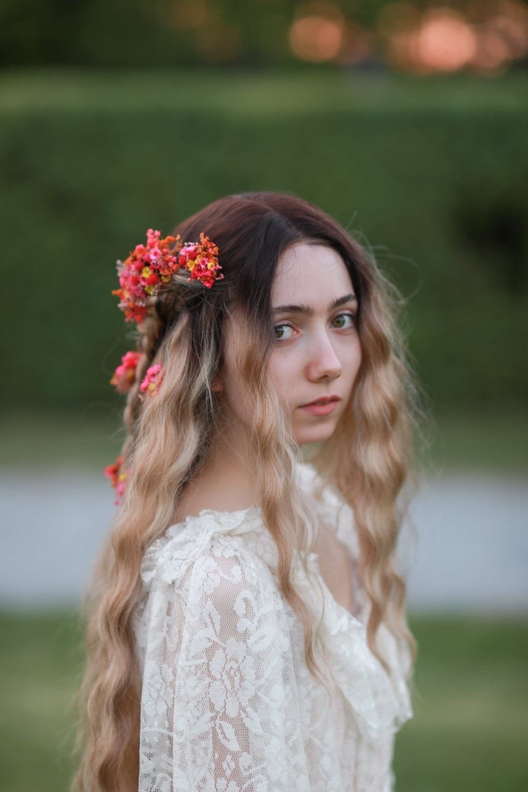 A young woman with long wavy blonde hair wearing a floral  comb and a white lace top, photographed outdoors against a blurred green background.