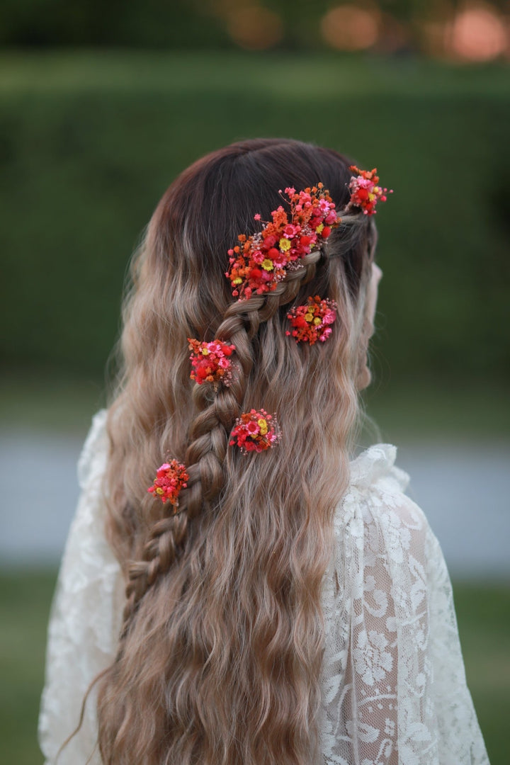 Long wavy blonde-to-brown ombre hair styled with a side braid and braided accessories, decorated with bright red and orange floral hair clips, shown from the back on a woman in a white lace top against a blurred green outdoor background.