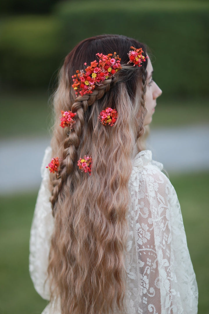 A woman shown from the back in profile, wearing a long wavy blonde-to-brown ombre hairstyle with braided details and multiple red and orange floral hair accessories. She is wearing a white lace top, set against a blurred green outdoor background.