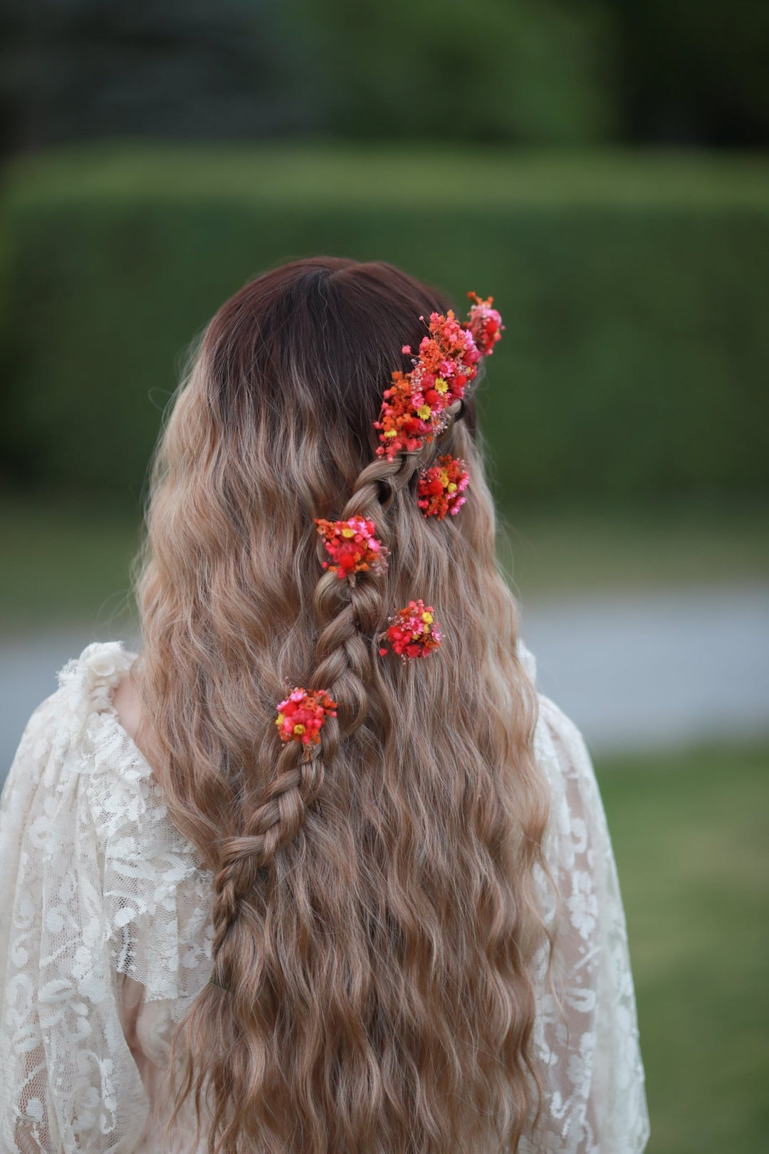 Long wavy blonde-to-brown ombre hair styled in a side braid adorned with small red and orange flowers, shown from behind on a model wearing a white lace top against a blurred green garden background.
