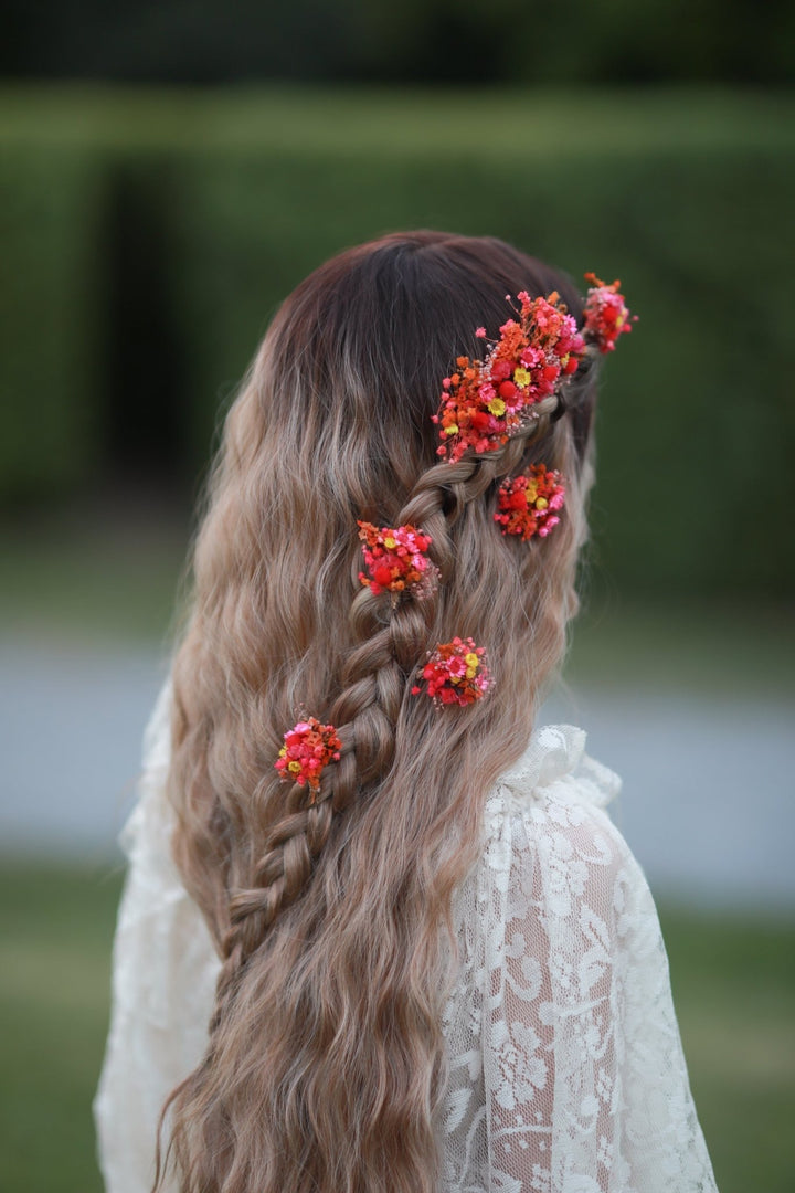 A woman seen from behind wearing a long, wavy blonde braid adorned with bright red and orange floral hair clips, set against a blurred green garden background.