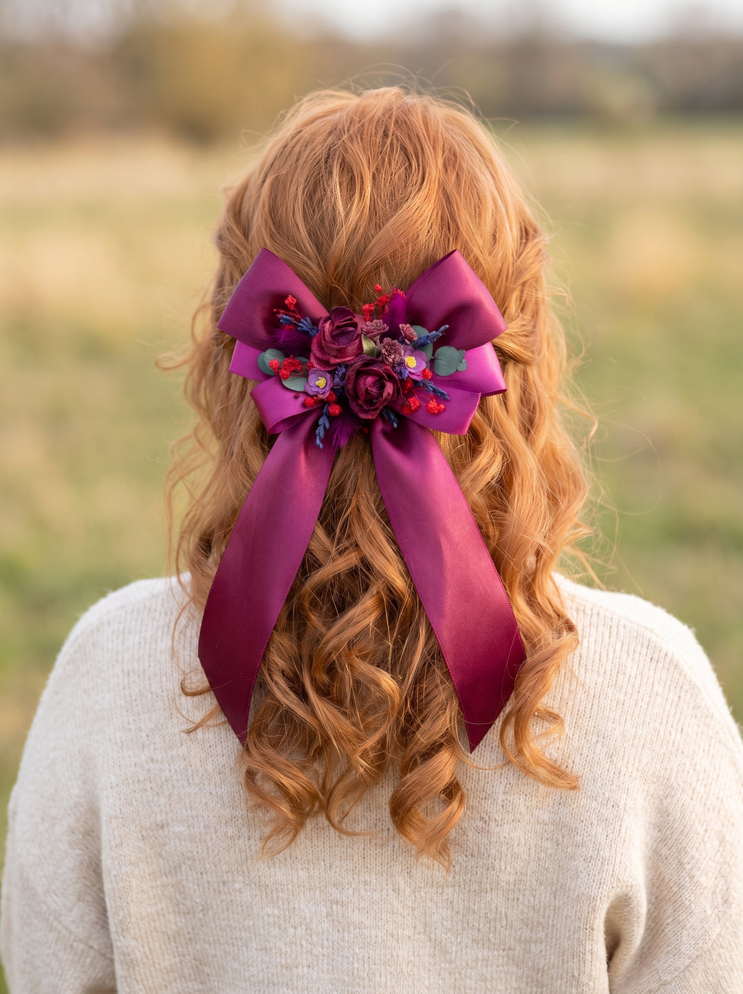 A person viewed from behind wearing a large magenta satin hair bow with floral embellishments in a grassy outdoor setting.