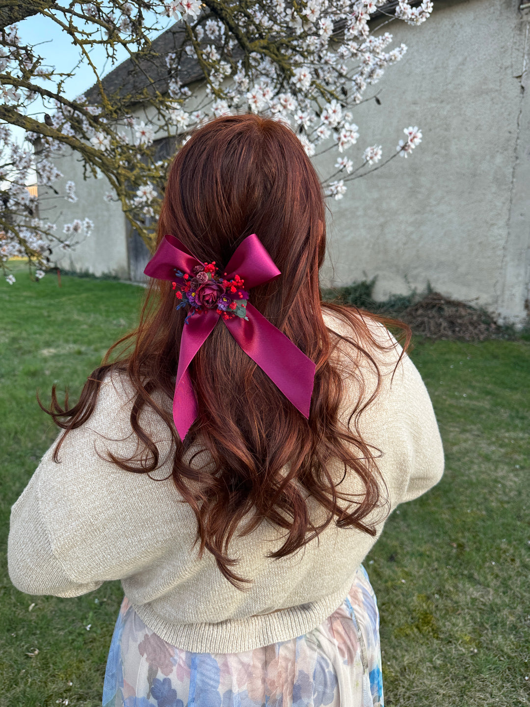 A person outdoors with long wavy brown hair, wearing a cream knit sweater and a floral skirt, with a bright pink ribbon and floral hair accessory tied in a low half-updo.