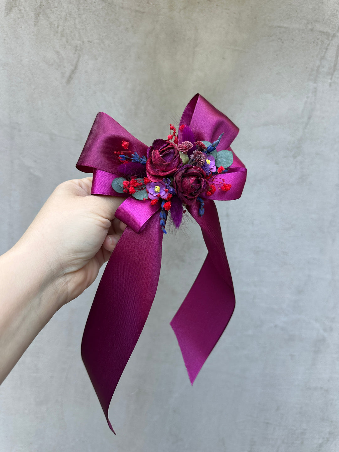 A hand holding a purple satin ribbon flower bouquet accessory with dried flowers and small colorful beads against a textured gray background.