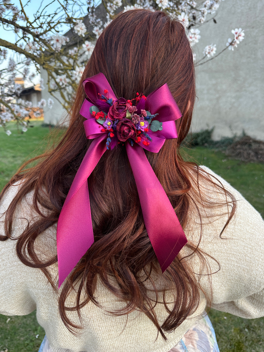 A brown wavy hair accessory featuring a large magenta satin ribbon bow with a colorful floral embellishment in the center, shown worn on a person's head outdoors.
