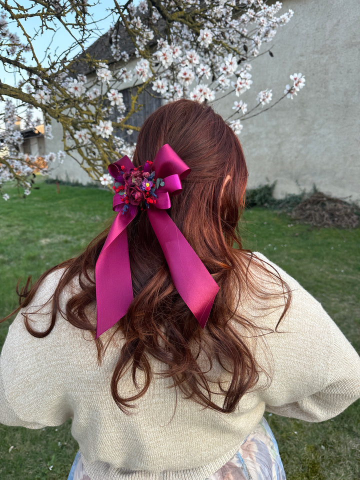 A person outdoors wearing a cream knit sweater, viewed from behind, with long wavy brown hair adorned with a large magenta ribbon hair accessory and floral embellishment.