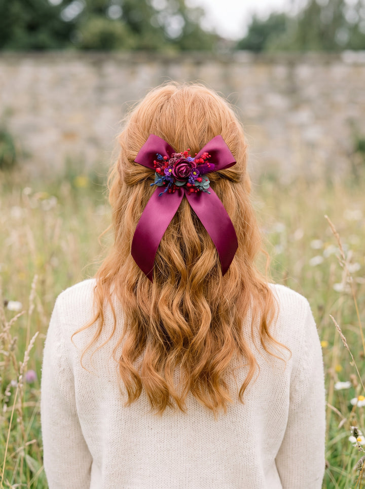 A woman viewed from behind in a grassy outdoor setting, wearing a cream knit sweater and a long wavy light brown hair accessory with a large deep purple ribbon bow and floral embellishment.