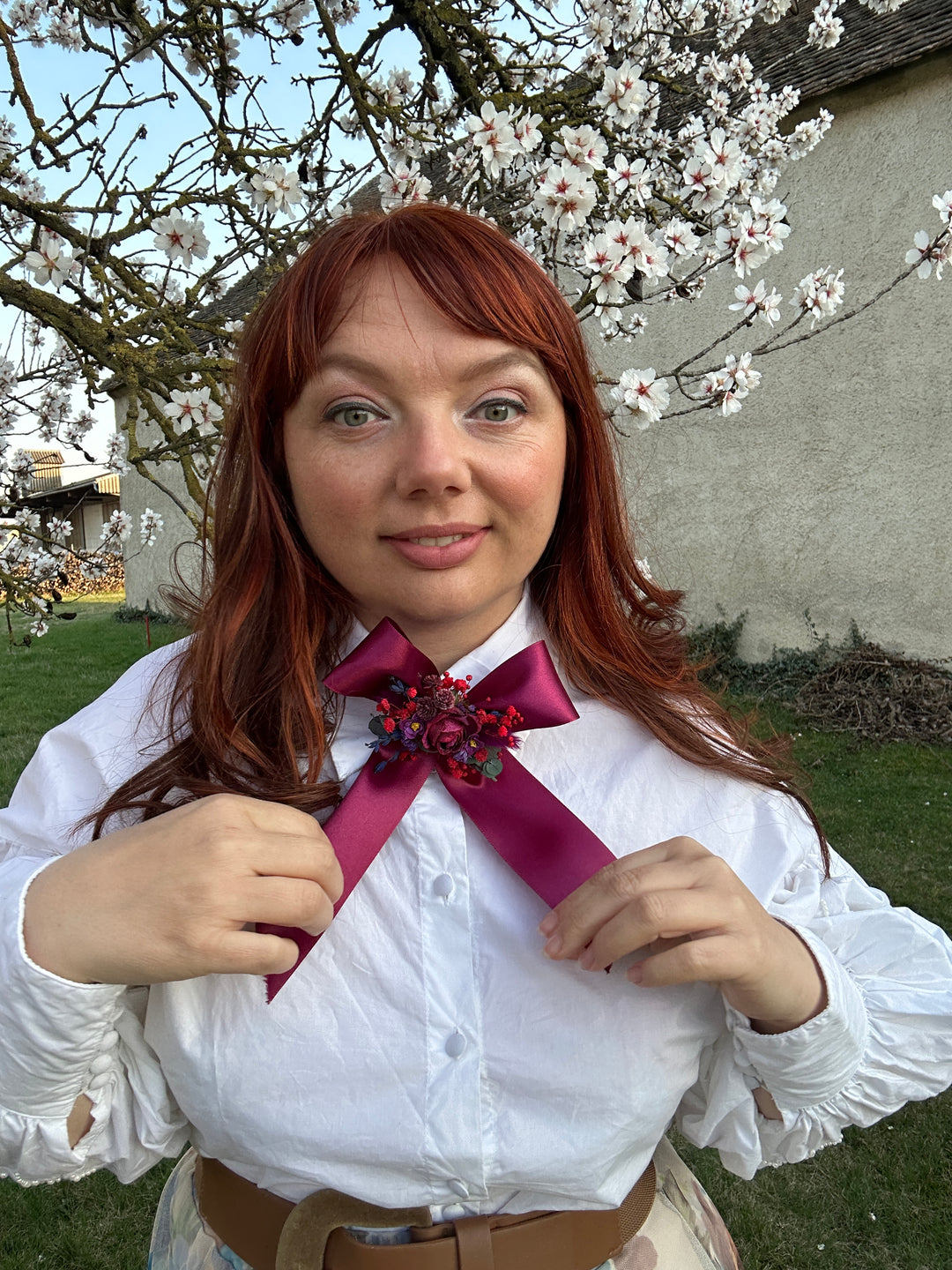 A woman outdoors wearing a white long-sleeve blouse with a large magenta satin bow and floral embellishment at the collar, paired with a brown belt. She is smiling and looking at the camera, with blooming white flowers and green grass in the background.