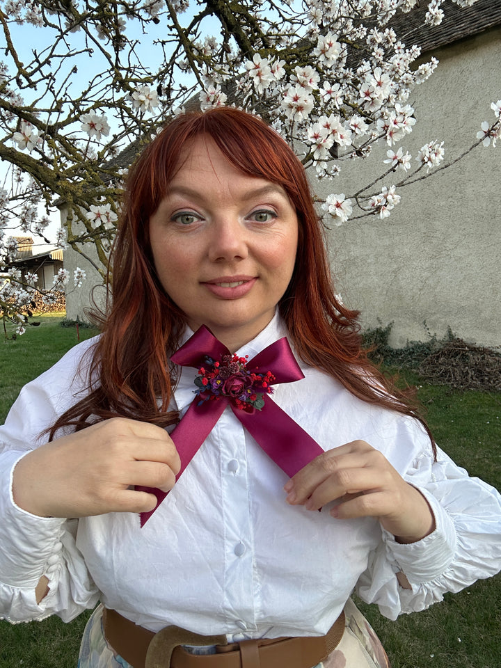 A woman outdoors wearing a white long-sleeve blouse with a large magenta satin bow and floral embellishment at the collar, paired with a brown belt. She is smiling and looking at the camera, with blooming white flowers and green grass in the background.