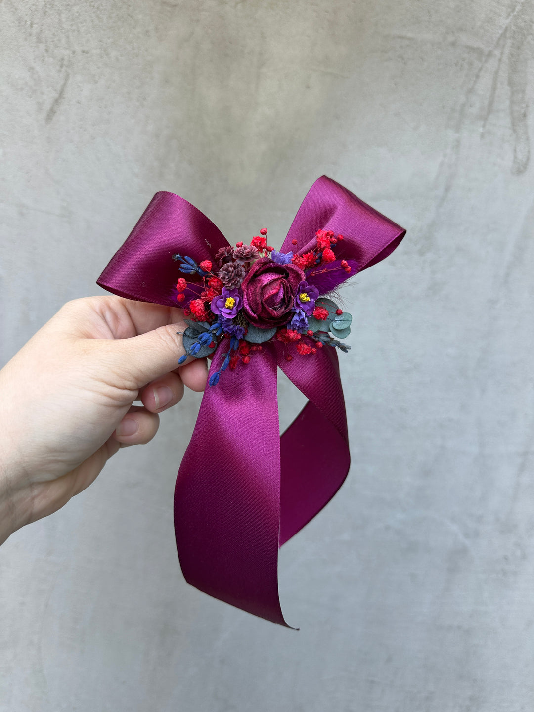 A hand holding a decorative hair accessory featuring a large magenta satin ribbon bow with a central floral embellishment in purple, red, and blue, set against a textured light gray background.