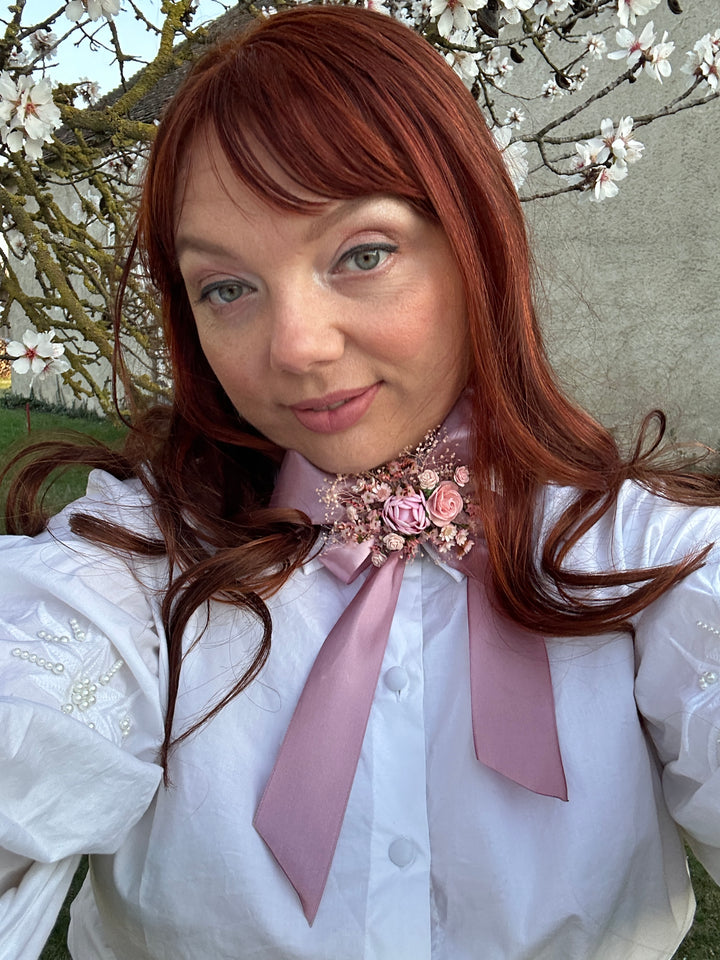 A woman with long auburn hair wearing a white blouse with a dusty pink ribbon and floral hair accessory, photographed outdoors among flowering branches.